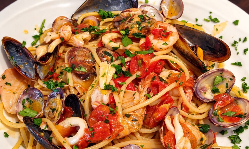 Pasta lunga con cozze, vongole, gamberi, calamari, pomodoro e prezzemolo, servita su un piatto bianco.