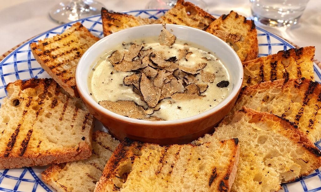 Ciotola di fonduta con lamelle di tartufo, circondata da fette di pane tostato su un piatto a quadri blu.