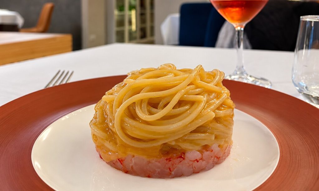 Nido di spaghetti su una base di tartare di gambero rosso, servito in un piatto con bordo marrone; sullo sfondo un calice.