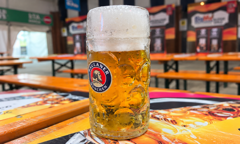 Boccale di birra Paulaner con schiuma e condensa, appoggiato sul tavolo con le panche dell’Oktoberfest Calabria sullo sfondo.