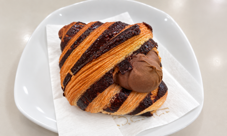Croissant con fasce dorate e scure e crema al cioccolato visibile sul lato, servito su un tovagliolo in un piatto bianco.