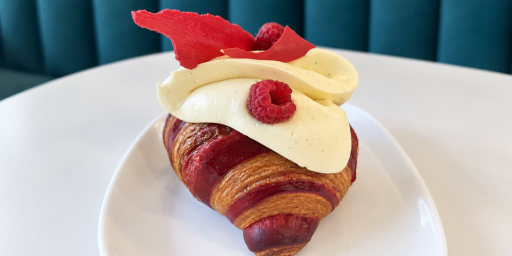 Croissant a fasce dorate e rosse, decorato con crema alla vaniglia, lamponi e cialde rosse, su un piatto bianco.