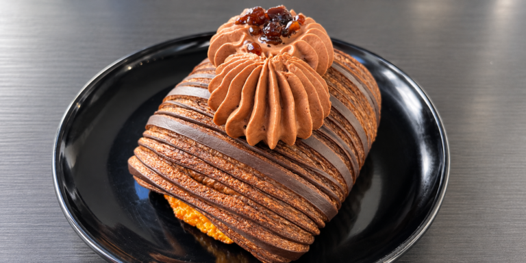 Pain Suisse con sfoglia al cacao, ciuffi di namelaka al cioccolato e sfere di caffè, su un piatto nero.