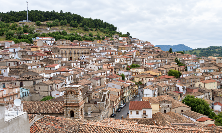 Panorama del centro storico di Cerchiara di Calabria, con case dai tetti in coppi, un campanile in primo piano e la collina alberata sullo sfondo.