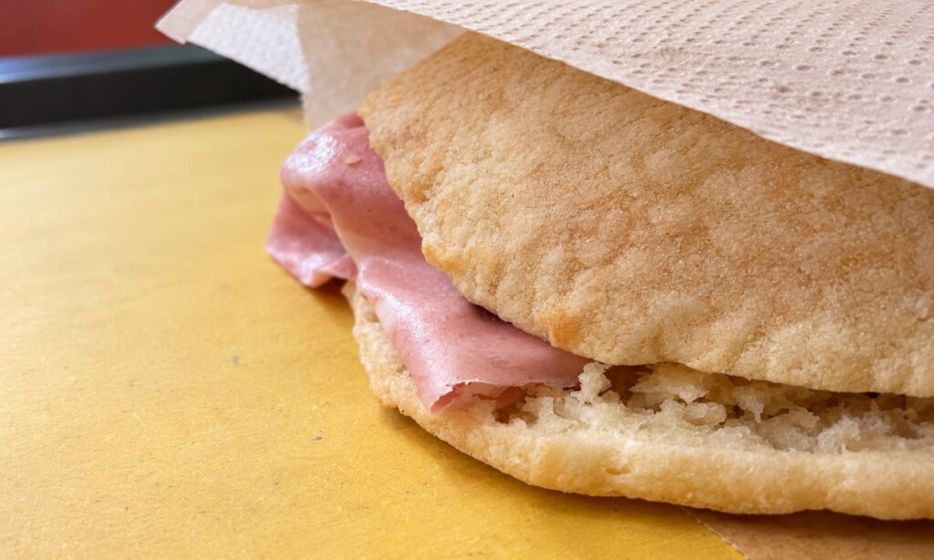 Primo piano della puccia con mortadella visibile tra le due parti del pane, appoggiata su un tavolo giallo.