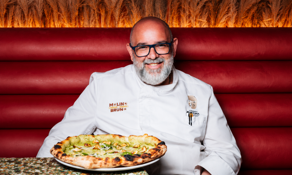 Lorenzo Fortuna sorridente, con occhiali e giacca bianca da pizzaiolo, seduto davanti a una pizza su un piatto, con una seduta rossa sullo sfondo
