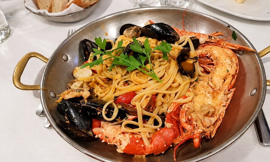 Pasta lunga allo scoglio con molluschi, crostacei e prezzemolo, servita in una padella metallica con due manici.