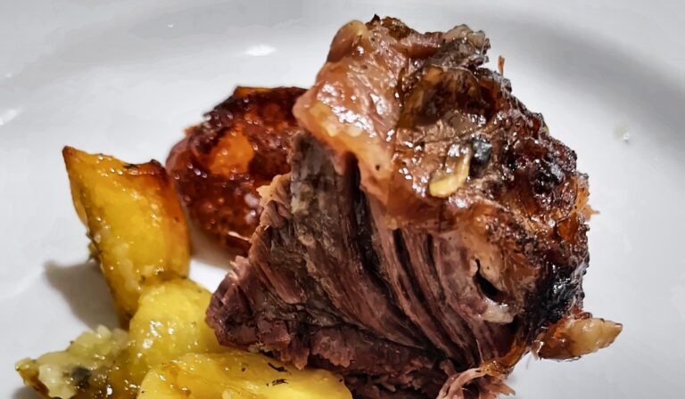 Primo piano di un pezzo di carne arrosto cotta a lungo, con la superficie scura e caramellata e le fibre sfilacciate, accanto a spicchi di patate al forno dorati e conditi con erbe aromatiche, su un piatto bianco.